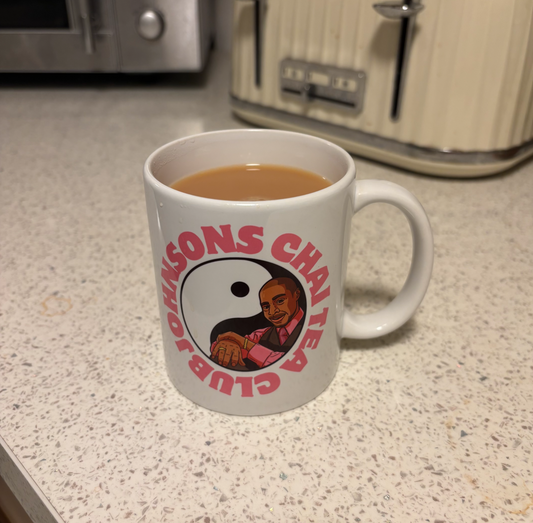 A white mug with a graphic design featuring a man's face and the text "JOHNSONS CHAI TEA CLUB" printed on it, placed on a kitchen counter.